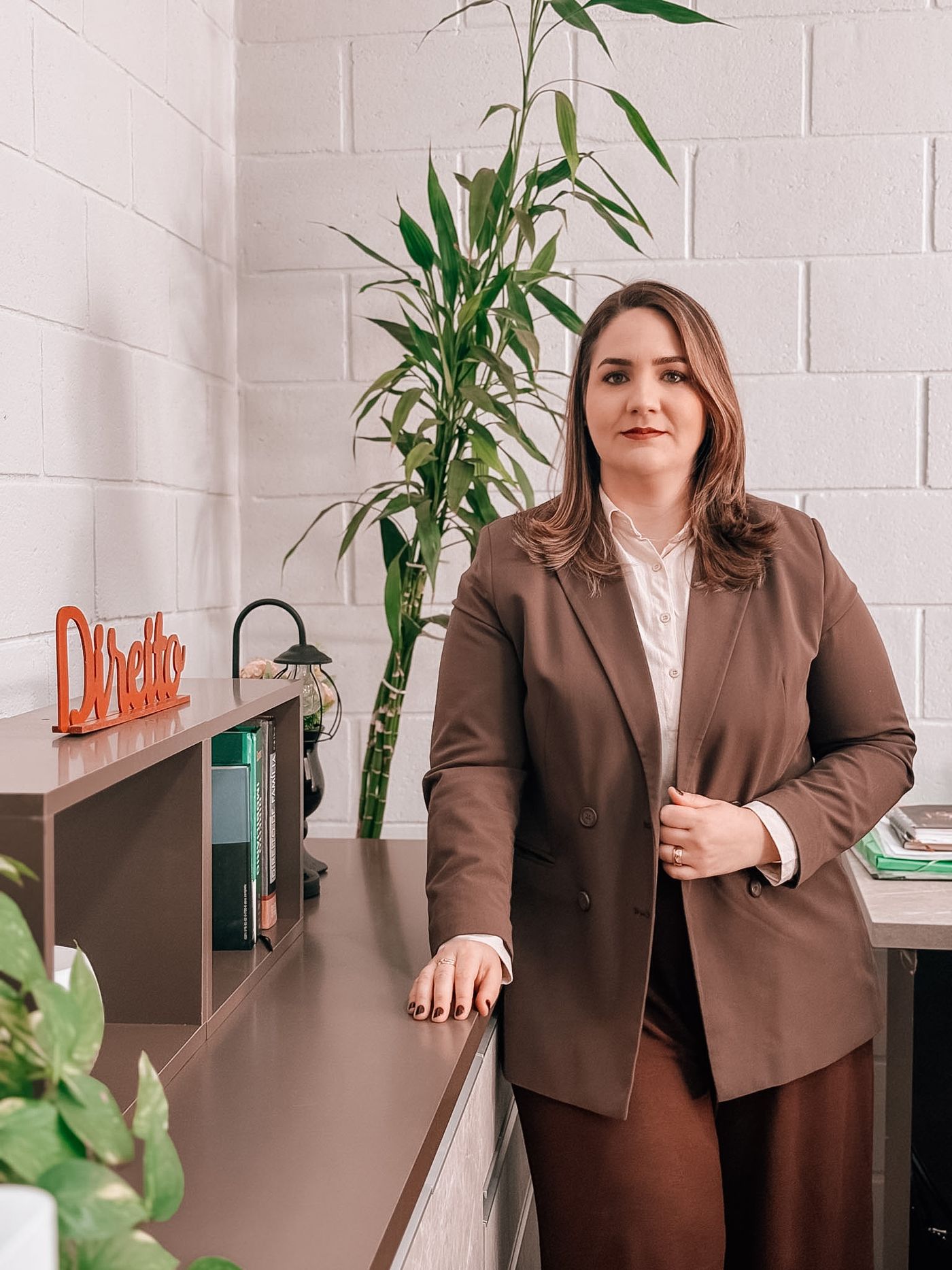 Advogada de blazer marrom no escritório, ao lado de uma estante com livros de direito e uma planta