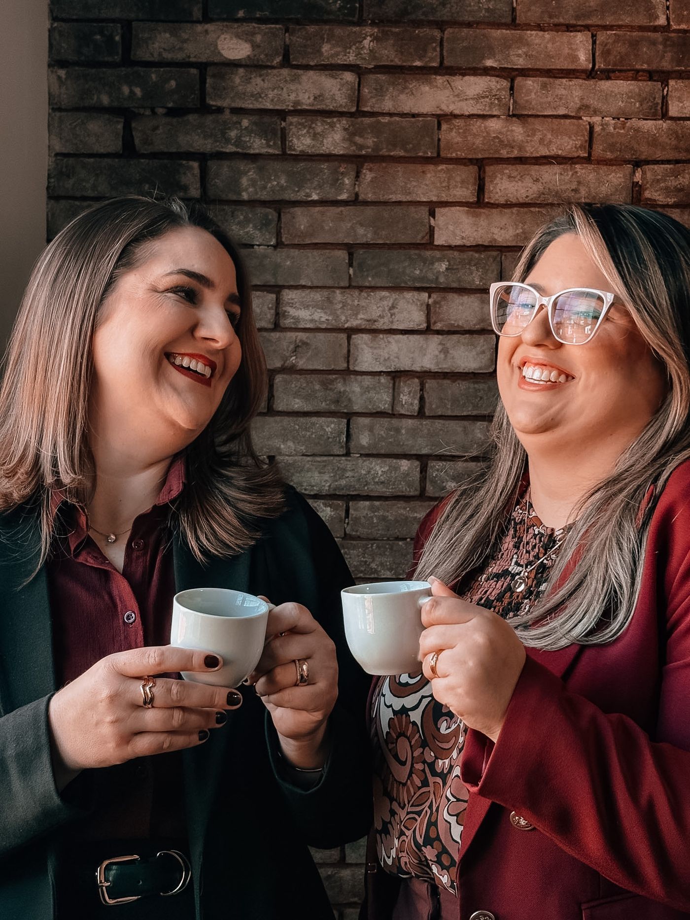 Duas advogadas do escritório sorrindo e tomando café diante de uma parede de tijolos